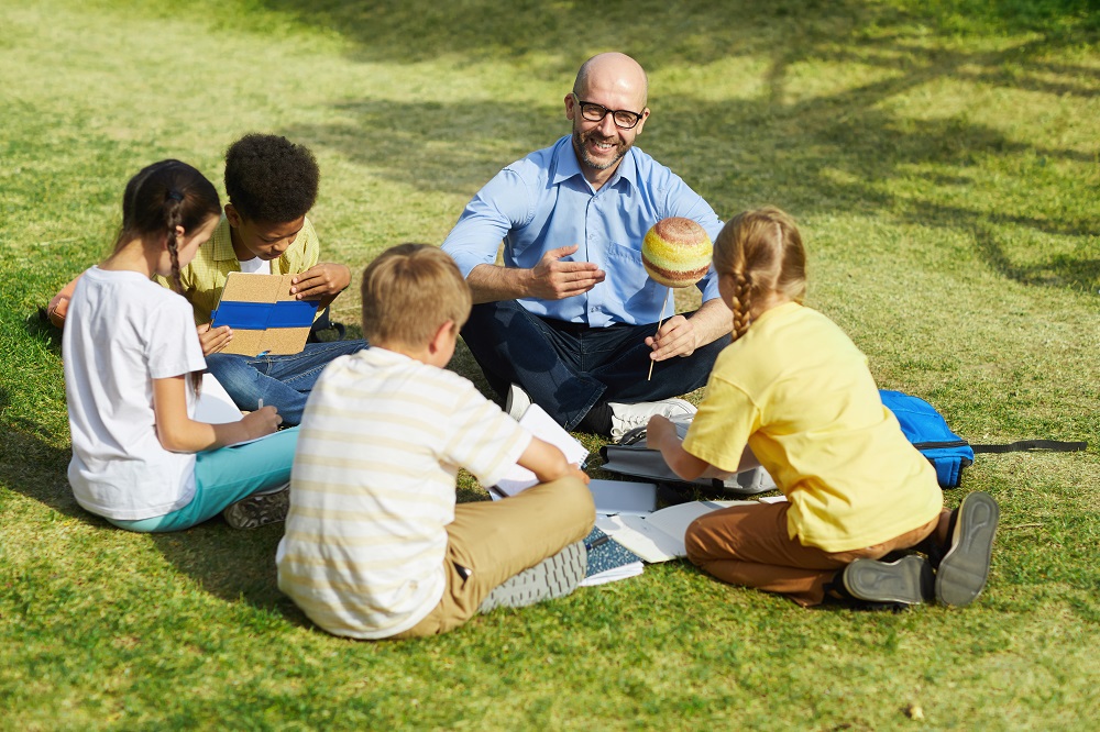 High angle portrait of bald male teacher pointing at planet model and smiling while talking to group of children during outdoor class in sunlight, copy space